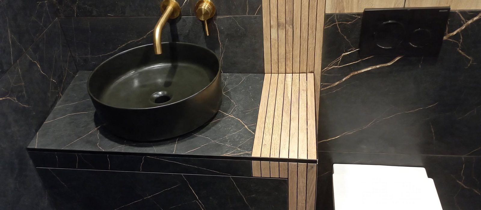 Afgewerkte badkamer in zwart marmer met zwarte wastafelkom, gouden wandkraan en eiken lattenwand
